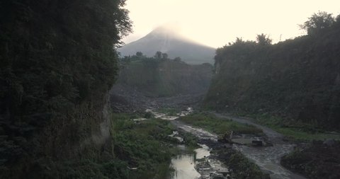 Reversing drone shot of eruption path with view of Merapi Volcano in sunrise time. The path is surrounded by trees. Bebeng River, Bego Pendem, Central Java, Indonesia