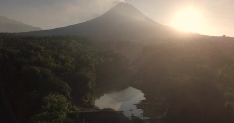 Reversing drone shot of eruption path with lake and view of Merapi Volcano in sunrise time. The path and lake are surrounded by trees. Bebeng River, Bego Pendem, Central Java, Indonesia