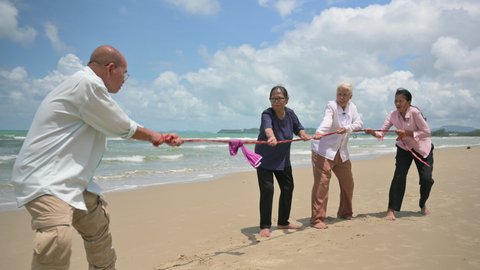 Happy Asian elderly tourist playing tug of war together and fun on beach in outdoors. Group of senior retirement and friends relax by pulling rope during tug of war game at coast in summer vacation. 
