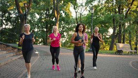 Fitness class for seniors. Group of active mature ladies with young black trainer running in urban park, practicing sport training, tracking shot