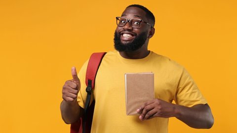 Excited young african american man 20s wears orange t-shirt backpack glasses hold book with blank screen workspace area show thumb up like gesture isolated on plain yellow background studio portrait