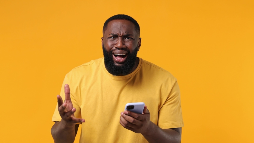 Sad irritated young african american man 20s wears orange t-shirt using mobile cell phone swear hears fake news unexpected rumor has some problems isolated on plain yellow background studio portrait | Shutterstock HD Video #1078443842