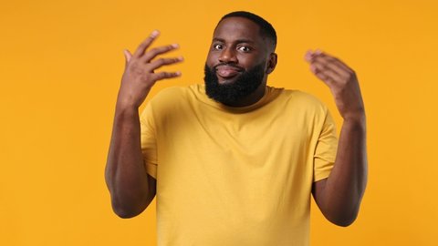 Fun confused shy puzzled young bearded african american man 20s wears orange t-shirt look camera spreading hands say oops ouch oh omg i am so sorry isolated on plain yellow background studio portrait