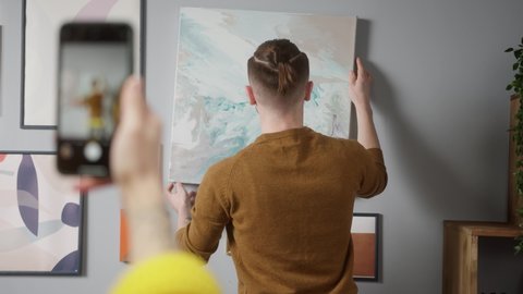 Young couple hanging picture on wall, woman making video on smartphone how man hangs framed picture in new apartments, move to real estate and new home.