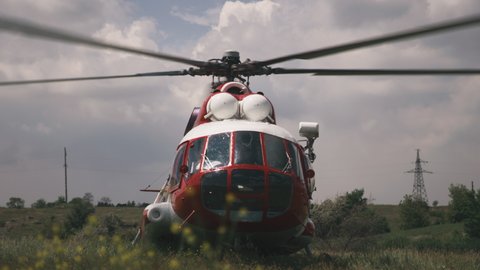 Red and white helicopter with rotating propeller landed in grassy field against cloudy sky during rescue operation in nature