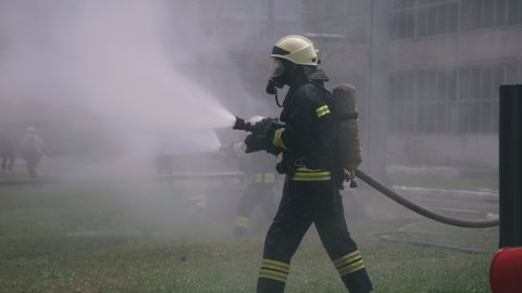 Firemen in protective uniform and respirators pouring jets of water from hoses while practicing to extinguish fire