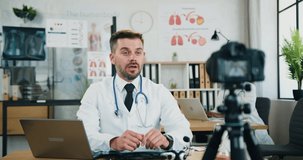 Attractive confident experienced bearded doctor sitting in front of camera in own medical office and record video for his internet audience