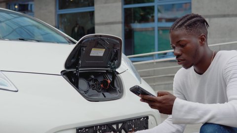 A young african american man connects an electric car to the charger and adjusts the process of charging the car battery using a cell phone