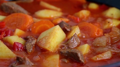 Beef stew with potatoes and carrots in tomato sauce in red pot, boils and gurgles, close-up.