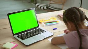 Home education for children. Little girl student with thin plaits writes in copybook near laptop with chromakey screen at table in room backside view