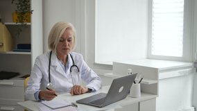 Doctor telehealth remote service. Skilled blonde mature therapist with stethoscope examines patient and makes notes at videochat via modern laptop in clinic