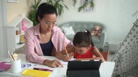 Online primary school education. Cute little Korean schoolgirl writes in copybook doing hometask with mommy nodding head during video lesson at home
