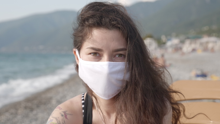 Half face portrait of a young woman with piercing gaze wearing protective mask on public beach. Concept of prevention against coronavirus. Health and safety life during pandemic, N1H1 Royalty-Free Stock Footage #1078474979