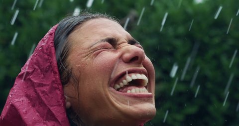 Authentic close up of carefree young woman wearing red protection cape is feeling free and smiling under the rain on a background of green trees. 