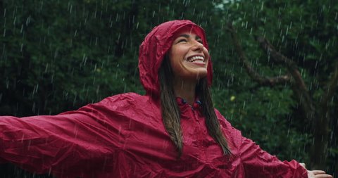 Authentic shot of carefree young woman wearing red protection cape is feeling free and smiling under the rain on a background of green trees. 