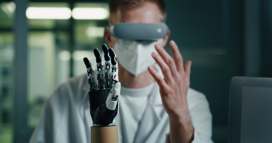 Young Biomedical Engineer is Wearing Virtual Reality Goggles and Controlling Remotely Bionic Limb Prosthesis. Professional Manipulating Artificial Palm. Robotic Innovation for People with Disability. Royalty-Free Stock Footage #1078476458