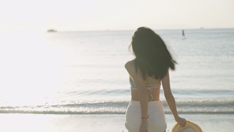 A rear view following on a sexy bikini girl holding hat and cheerfully running into sea water, walking against sunlight, hot beautiful summer vacation, a long straight black hair flowing in the air