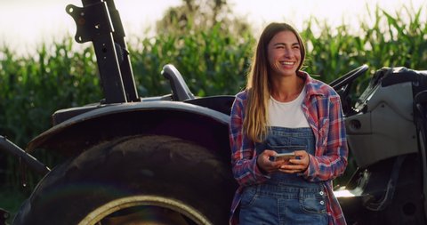 Cinematic shot of female farmer is using smartphone and smiling satisfied with her work on countryside farmland corn fields. 
