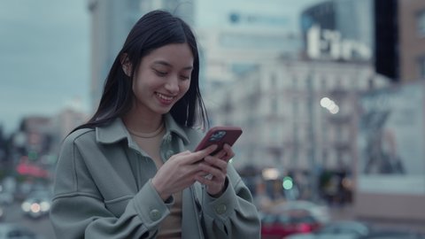 Happy japanese woman in casual outfit standing on city street with cell phone in hands. Charming brunette chatting with friends online using modern smartphone.
