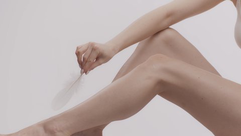 Modern hair removal. Close up shot of unrecognizable caucasian lady caressing smooth skin of her perfect legs with feather, white studio background, side view, slow motion