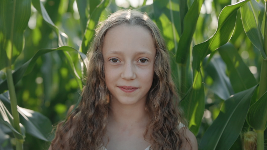 Pleasant school girl in casual clothes smiling sincerely while standing at corn field. Pretty face of caucasian child. Rustic lifestyles.