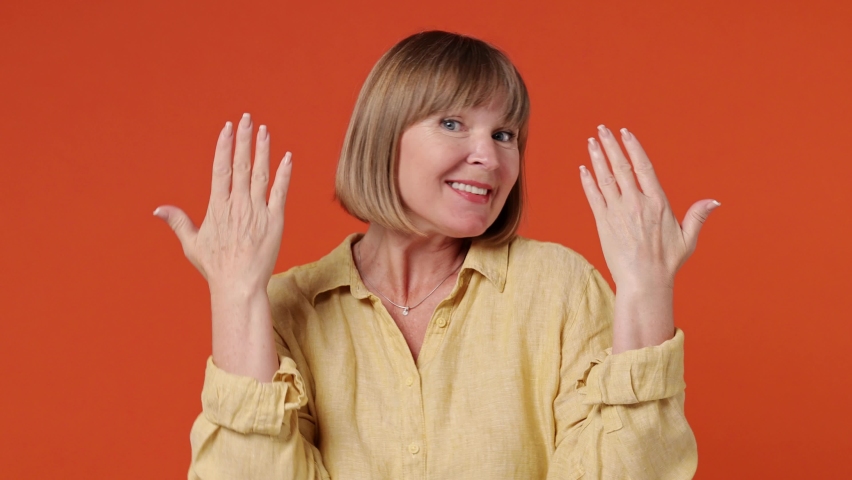 Mature senior elegant lady woman 55 years old wears yellow shirt dance clenching fists waving rising expressive gesticulating hands have fun enjoy celebrate isolated on plain orange background studio