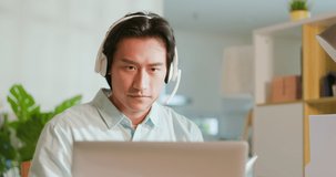 asian man has video meeting - he wearing headset is using laptop and taking documents at home