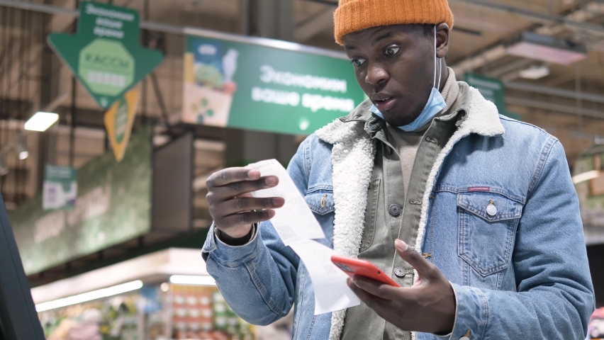 African-American man in a medical mask looks at a check at the checkout counter in a supermarket with a phone in his hands and is surprised and indignant at the expensive products. Concept of the rise Royalty-Free Stock Footage #1078521239