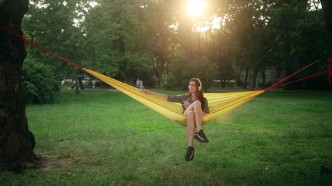 Young Cheerful red-haired woman resting on hammock. Pretty lady enjoys listening chill music. Beautiful girl wear headphones having fun