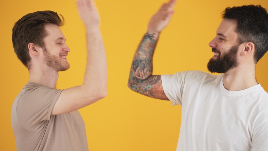 Two positive friends guys are giving five standing isolated over yellow wall in the studio