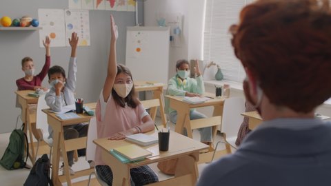 Handheld shot of group of middle school children in face masks sitting at desks in classroom and raising hands to answer question by female teacher