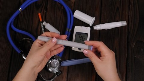 A diabetic woman dials the right dose of insulin on an insulin pen sitting at a wooden table, a stethoscope, a glucose meter and lancets lie nearby. Diabetes and prevention of diabetes complications.