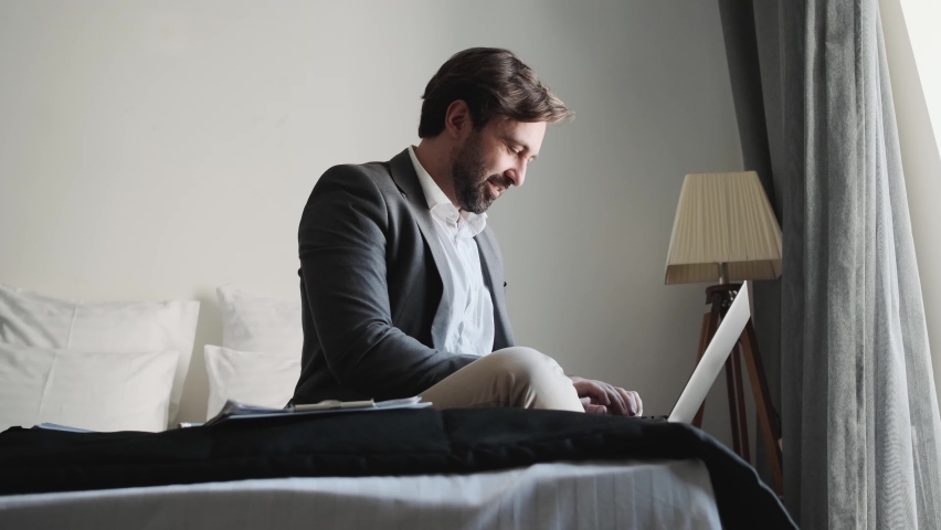 The side view of the smiling man in an office suit typing something on the laptop in the apartment