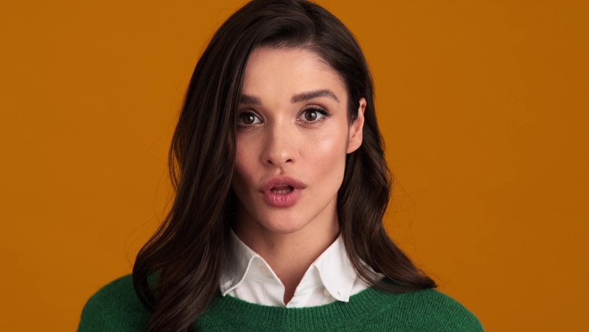 A close-up view of the positively surprised woman in a green sweater and shirt saying wow in an orange studio | Shutterstock HD Video #1078528025