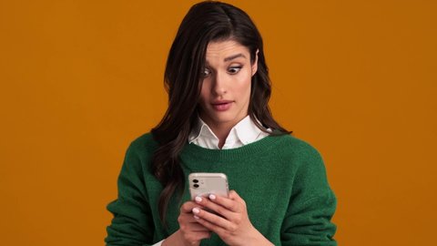 An angry woman in a green sweater and shirt typing something on the phone and raising an eyebrow while standing in an orange studio