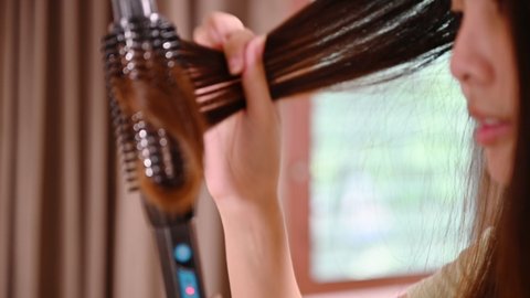 Asian woman curling hair with hair curler fashion with herself in front of mirror in bedroom