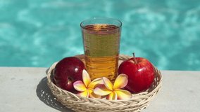 Video footage of glass with apple juice, bamboo straw, red apple, tropical flower frangipani and bubbling blue swimming pool on background.
