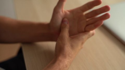 Close-up hands of unrecognizable suffering young man massaging painful palm sitting at desk with laptop in home office. Male massaging painful hand from symptoms of Peripheral Neuropathy.