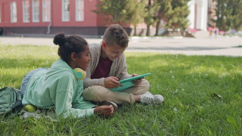 Medium shot with slowmo of cheerful African-American 11-year-old girl relaxing on green grass in schoolyard and chatting with 13-year-old boy playing on tablet