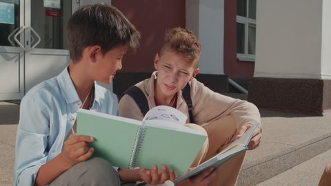 Handheld shot of 12-year-old and 13-year-old boys sitting on steps of middle school building and comparing their homework answers