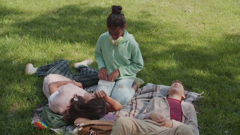 Handheld tracking shot of schoolkids relaxing on blanket laid out on green grass in park on summer day. Girl playing with sensory fidget toy