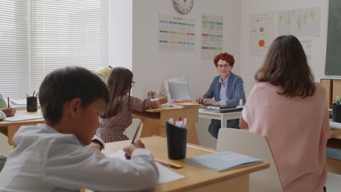 Handheld tracking shot of middle school children sitting at their desks and writing in notebooks while cheerful female teacher talking and answering questions