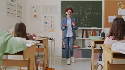 Handheld tracking of cheerful female math teacher standing by blackboard and explaining information to group of middle school students