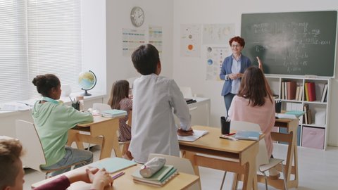 Handheld tracking shot of group of middle schoolers sitting at desks in classroom and raising their hands to answer question. Cheerful female teacher asking boy to answer it