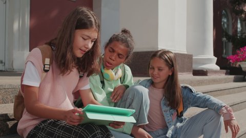 Handheld tracking of three girls sitting on school steps and chatting while using tablet