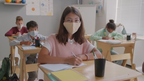 Handheld portrait of 13-year-old girl in face masks sitting in classroom with other students and writing in notebook during lesson, then looking at camera