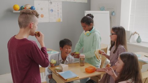 Medium shot of happy schoolchildren chatting and laughing while eating sandwiches and fruit in classroom during break
