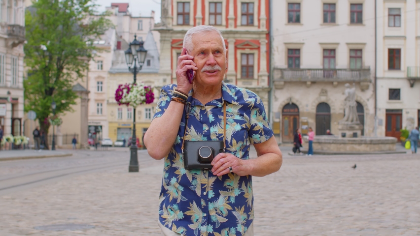 Excited elderly old tourist man talking on mobile phone while walking on city street of Lviv, Ukraine. Senior stylish mature grandfather enjoying smartphone conversation and traveling. Urban concept