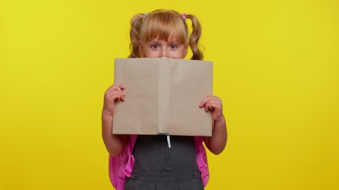 Attractive blond teenage student girl kid in school uniform wears pink backpack peeping while hiding behind a book, isolated on yellow studio background. Funny pupil. Education, back to school, study