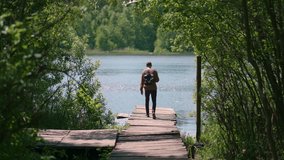 A young man with a backpack walks along the pier on the shore of the lake in the forest, shoots a walk and picturesque views of nature on a mobile phone. The tourist enjoys the beautiful scenery